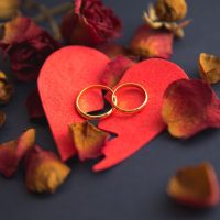 Gold wedding rings resting on a broken red heart surrounded by dried rose petals, symbolizing the emotional impact of divorce and relationship separation.