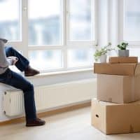 WomanMoverWithCardboardBoxesMovingInOutCityApartment Person sitting on a windowsill with one leg raised, surrounded by moving boxes in a bright room, symbolizing transitions related to family law matters like divorce and custody.