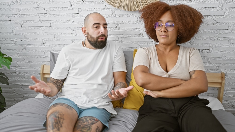 UpsetBeautifulCoupleSittingTogetherOnBedInBedroomArguing